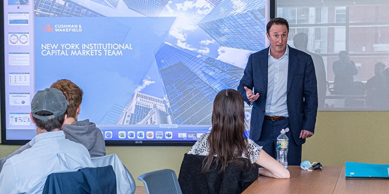 A man speaking to a class of students with a powerpoint 
