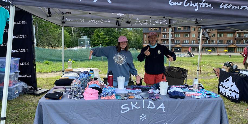 Two women stand selling ski hat products 