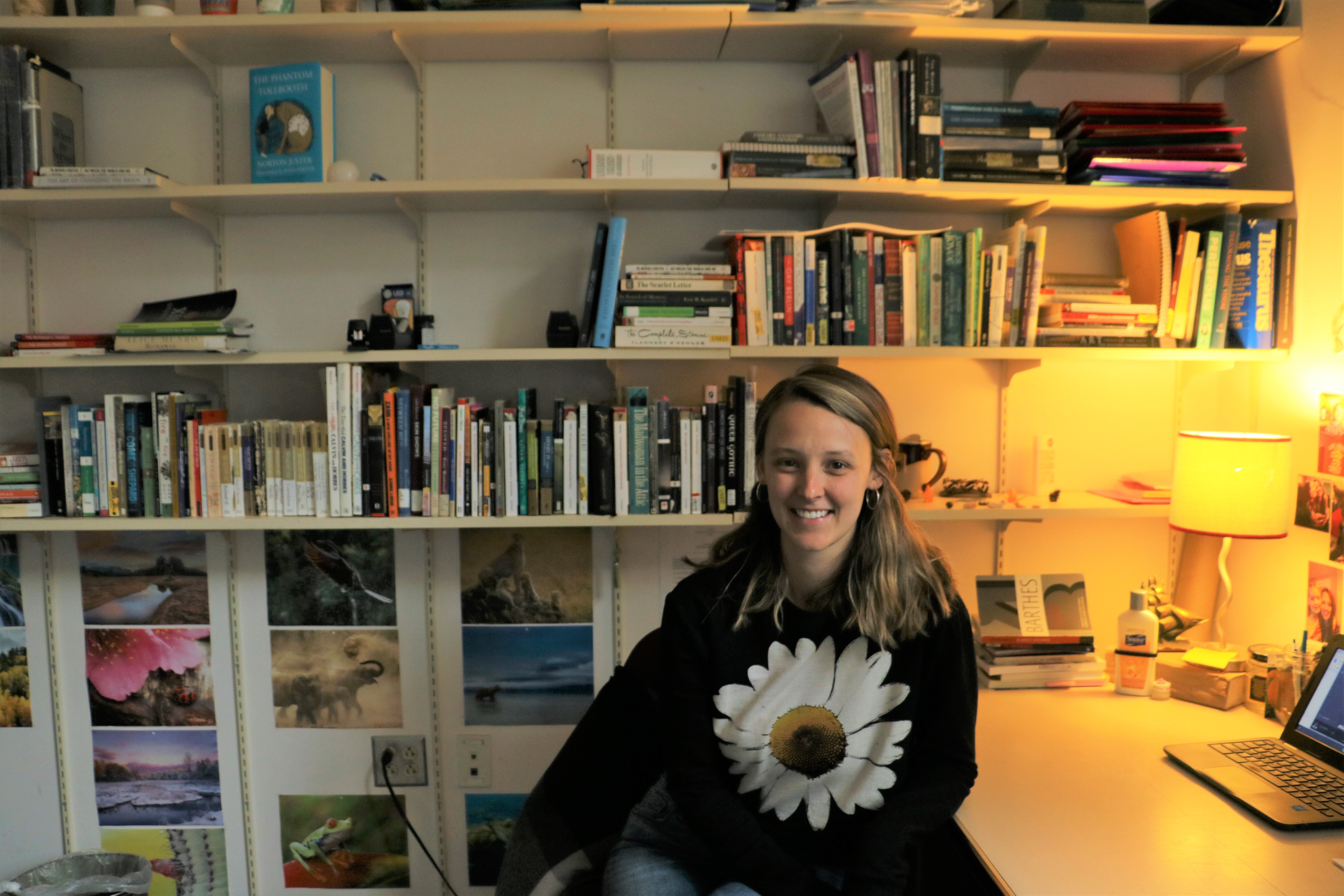 Casey O'Reilly, shoulder length light brown hair, smiling at the camera and sitting at her desk in front of a bookcase