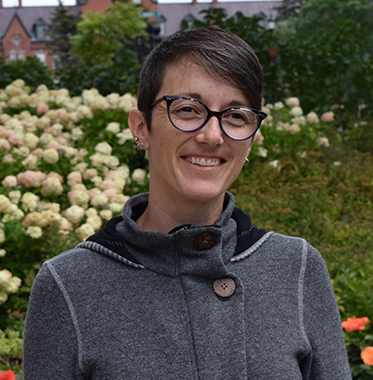 Headshot of Kate Jerman wearing grey sweater in front of flowering trees with Old Mill in background