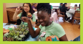 Woman wearing green shirt holding planting pottery and paint brush.