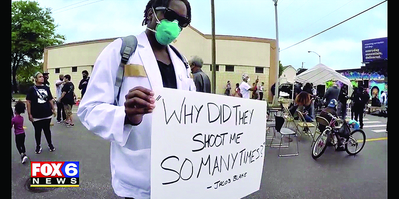 TV screen view of Christopher Veal in white coat at a protest carrying a sign stating 'Why did they shoot me so many times?"