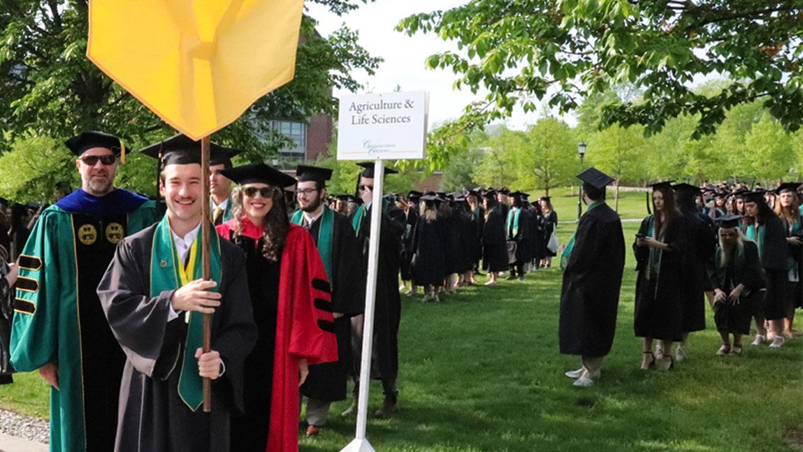 A UVM CALS college student is holding a yellow banner while graduating students line up behind him.