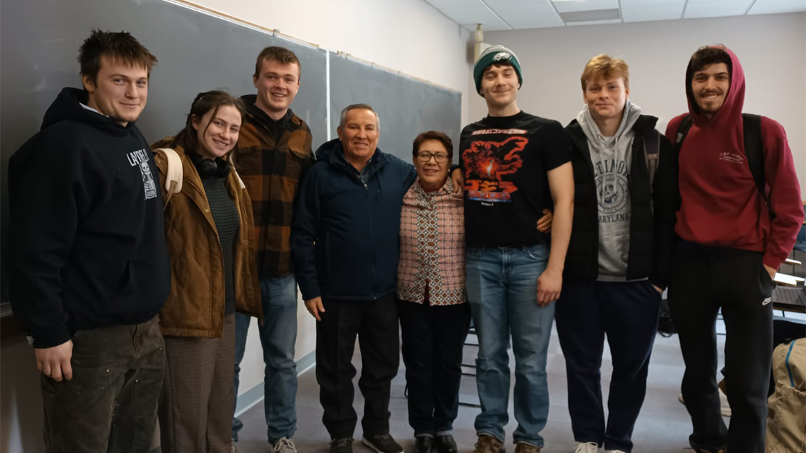 Students standing in a group indoors with a blackboard behind them. 