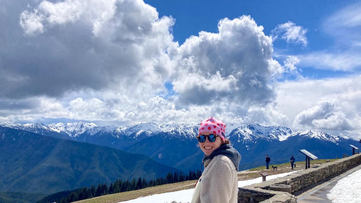 Person outdoors wearing a sweater and hat looking over a mountain view with puffy clouds in a bright blue sky