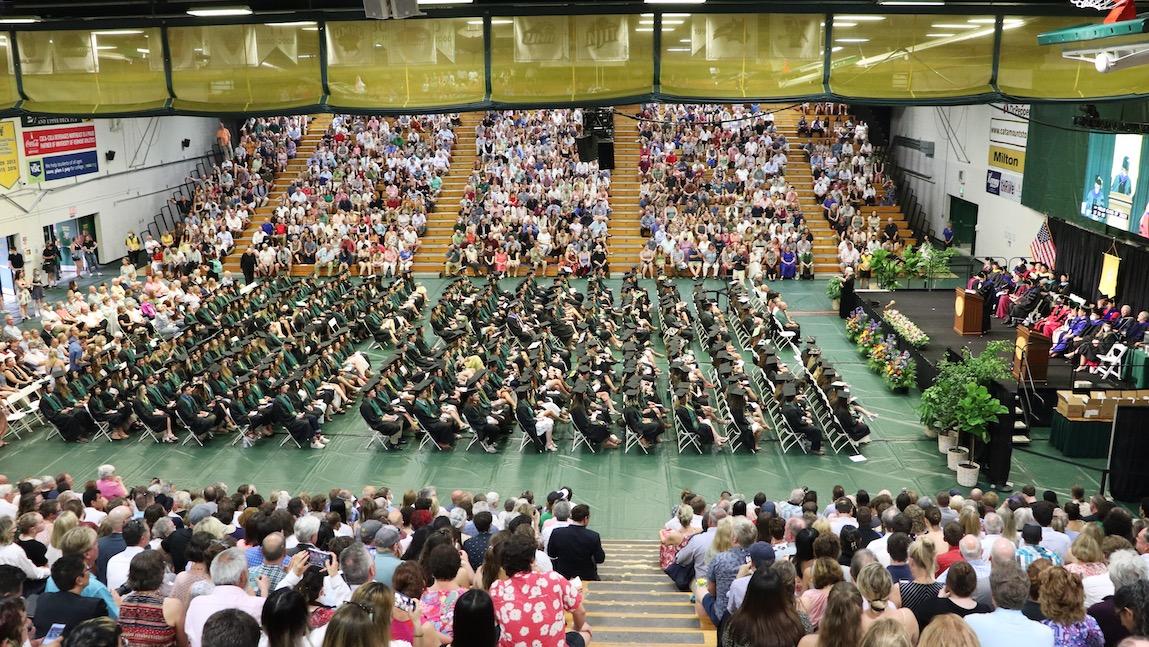 wide angle photo of commencement ceremony; families in bleachers of gymnasium with students seated on the gymnasium floor toward the commencement platform.