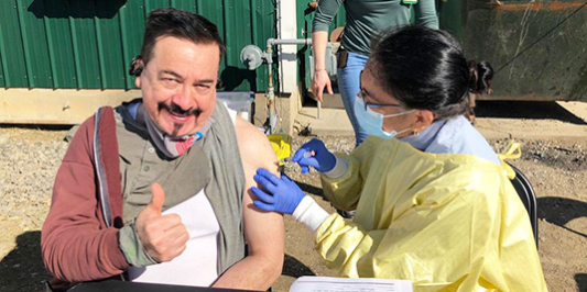 Alejandro giving a thumbs-up getting vaccinated by a health worker in protective gear