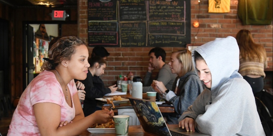 A small group of students sitting in a row of tables at the Skinny Pancake on campus. The two students in the front facing each other appear most visible – the one on the left is wearing a faded red tie-and-dye shirt and eating a meal. The student on the right is wearing a grey hooded sweatshirt, and smiling while working on their laptop. 