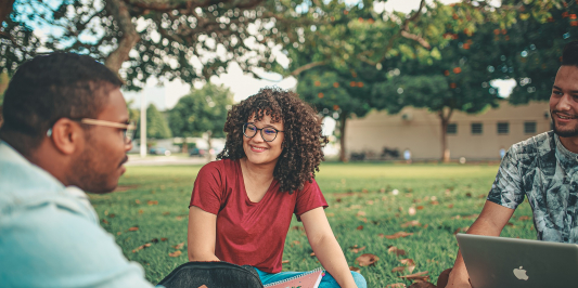 A group of young people sit together on a patch of grass on a University campus. A young woman of color, with loose, curly hair and glasses, listens attentively to a Black man seated to her right. He has short natural hair and glasses and is paused mid-utterance.