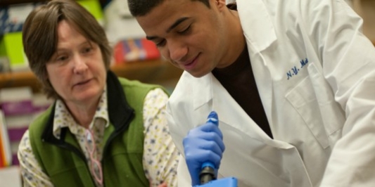 A student in a lab coat conducts an experiment while his faculty mentor looks on