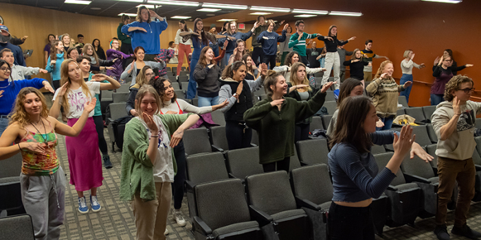 Students in a lecture hall follow along in a sequence of dance moves led by professor Paul Besaw.