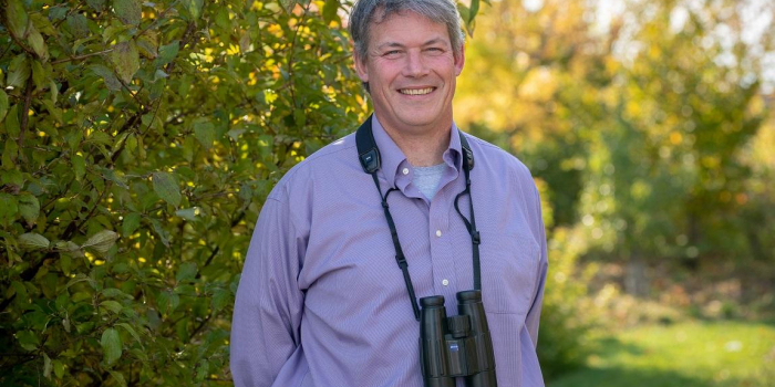 Allan Strong outside in front of greenery with binoculars around his neck.