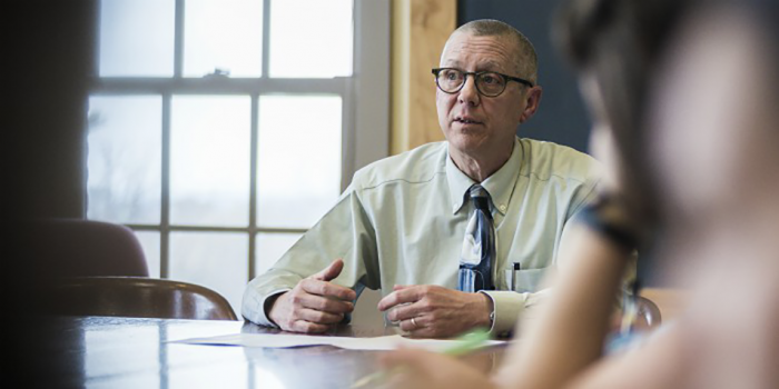 Bob Taylor seated at a table talking with others with light streaming in a window behind him