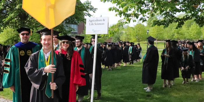 A UVM CALS college student is holding a yellow banner while graduating students line up behind him.
