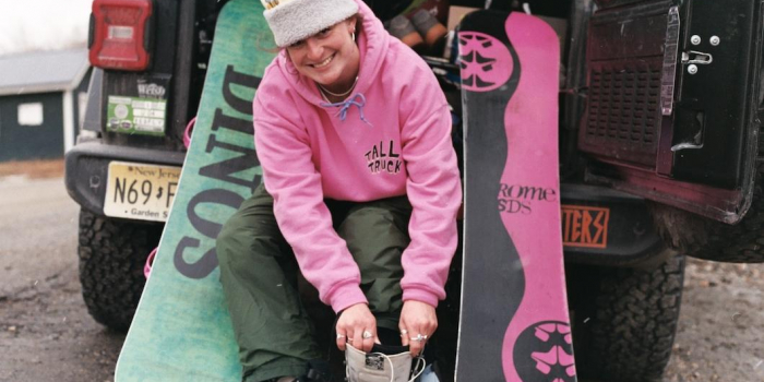woman wearing a hat sitting on hatchback of car putting on snowboard boots with two snowboards resting on either side of her.