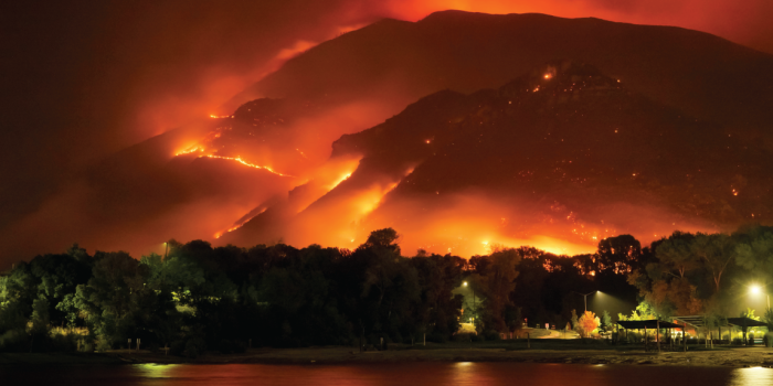 Image shows a forest fire in the background on mountains with a city and pond in the forefront. 