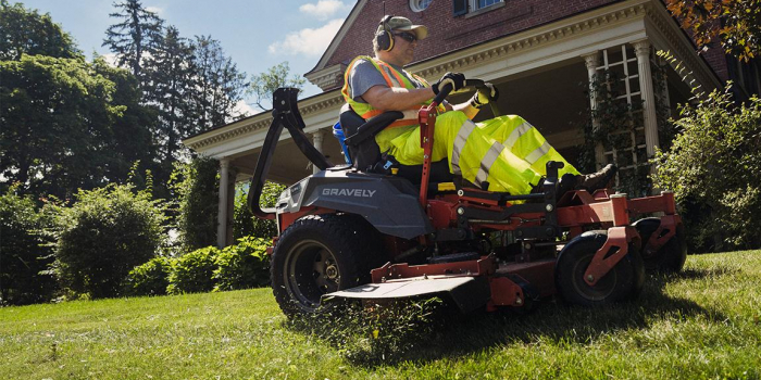 A man wearing a brightly colored safety vest operates a ride-on lawnmower in front of a building on UVM's campus