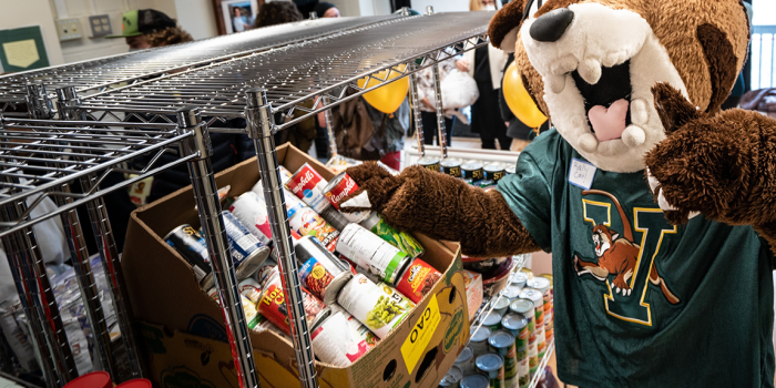 Rally Cat poses for the camera in front of shelves of non-perishable food items in UVM's student food pantry.