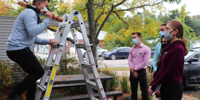 Person climbing a ladder while holding a long object over their shoulder, and three other people watching.