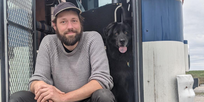 Serge Wiltshire. A man with dark hair and a beard in a grey sweater and hat sits with a black dog. 