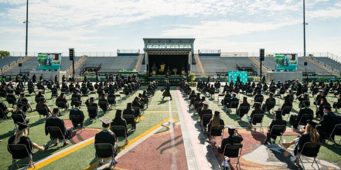 graduates seated in chairs 6 feet apart on the University of Vermont field with commencement stage in the background