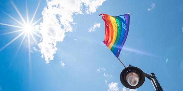 Rainbow pride flag flies off of lampost against blue sky with bright sun