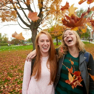 Two UVM students under a tree with fall colors in the branches and on the ground