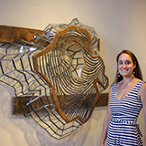 Artist Stefania Urist standing next to one of her wood and carboard creations hung on a wall.