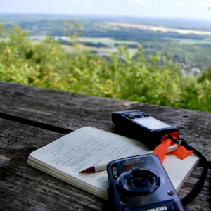 a notebook, camera, and GPS lie on a picnic table with a beautiful vista beyond