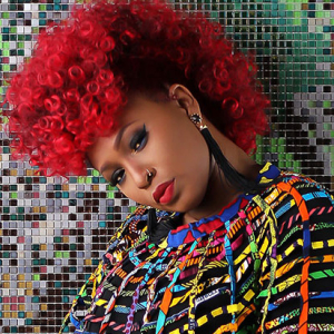 Mumu Fresh with red hair posing in front of a colorful mosaic wall