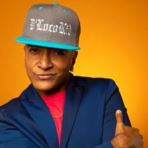 D'Lo in front of an orange background wearing a grey and blue baseball cap that has "D'Loco Kid" embroidered on the front