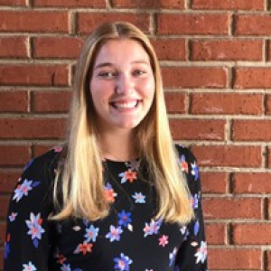 Alexandra smiling and standing in front of a bright red, brick wall
