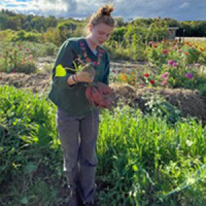Evie standing in a field of blooming flowers with her arms overflowing with produce