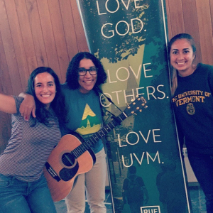 Three students standing beside a green banner that says "Love God. Love UVM."