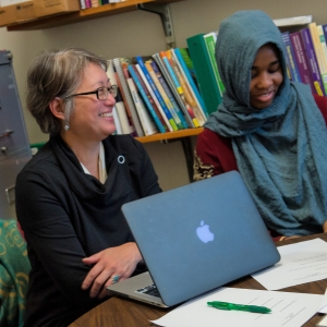 Professor, wearing glasses, sitting at table with student laughing.