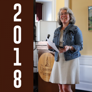 2018: Sheila Boland Chira, wearing a denim jacket over a gray dress, stands in front of a podium with a smile.