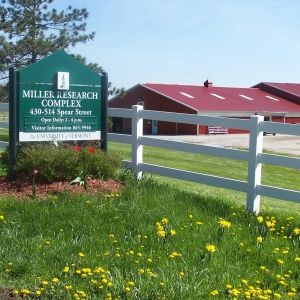 Miller Research Farm sign of the Miller Research and Education Center with a white fence and red farm buildings in the background.