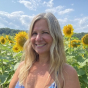 A white woman in her mid-forties, with loose blonde hair falling to her shoulders, stands smiling confidently in a field of sunflowers, under a pale and summery sky.