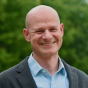 A bald white man in his early forties, wearing an open-collared pale blue shirt under a gray blazer, grins openly at the camera.