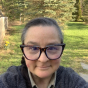 A pale-skinned non-binary person, wearing dark hair in a braid over one shoulder and large, heavy-framed glasses, stares directly into the camera with the hint of a smile.