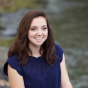 India Hoover. A pale-skinned woman in her twenties, with shoulder-length, wavy auburn hair, smiles broadly at a camera while sitting next to a creek.