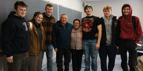 Students standing in a group indoors with a blackboard behind them. 