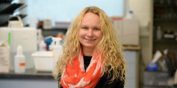 Jana Kraft. A person with long blonde hairs in an orange scarf sits with a laboratory as the backdrop. 