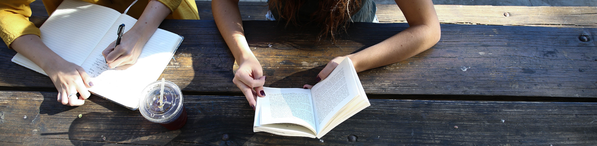 the hands of two women reading and writing at an outdoor table